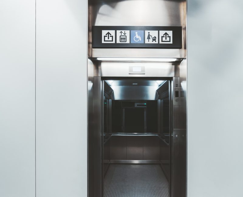 A modern lift inside of an airport Vertical view of a modern chrome elevator with opened doors indoors of a shopping mall, an airport terminal or a train station depot: multiple signs above the doors, the mirror inside